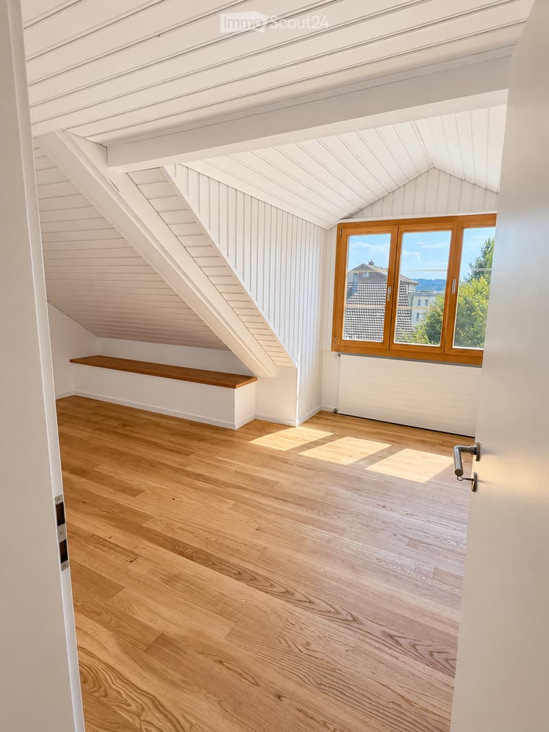 Bright, empty attic room with wooden flooring, white paneled walls, and a skylight window offering a view of the neighborhood.