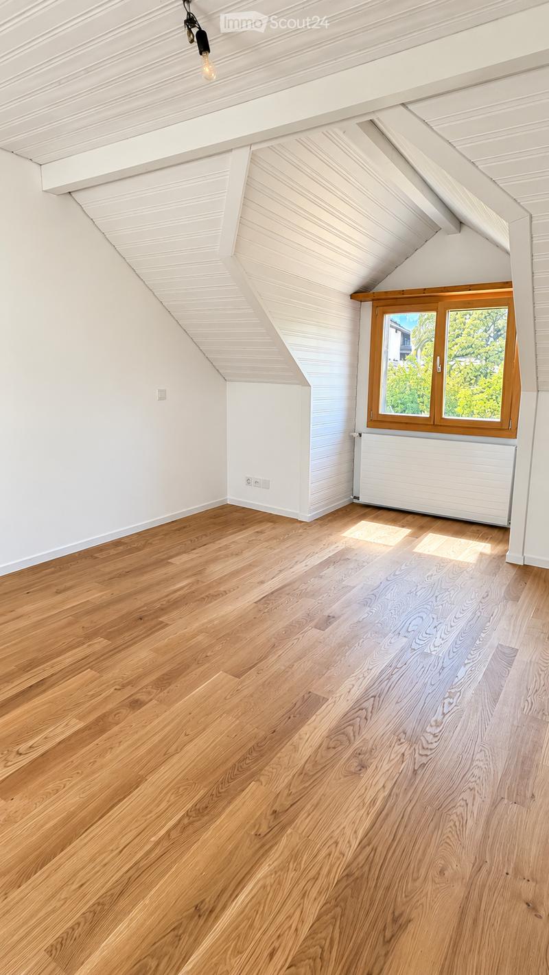 Empty attic room with wooden flooring, white walls, and a slanted ceiling. Features a window with wooden frame and radiator below it.