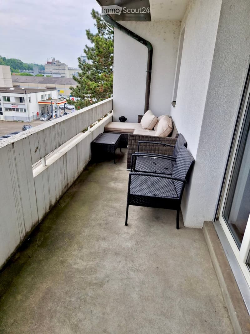 The image shows a balcony or terrace area with a wicker furniture set, including a sofa and chairs. The balcony overlooks a cityscape with buildings and trees visible in the background.