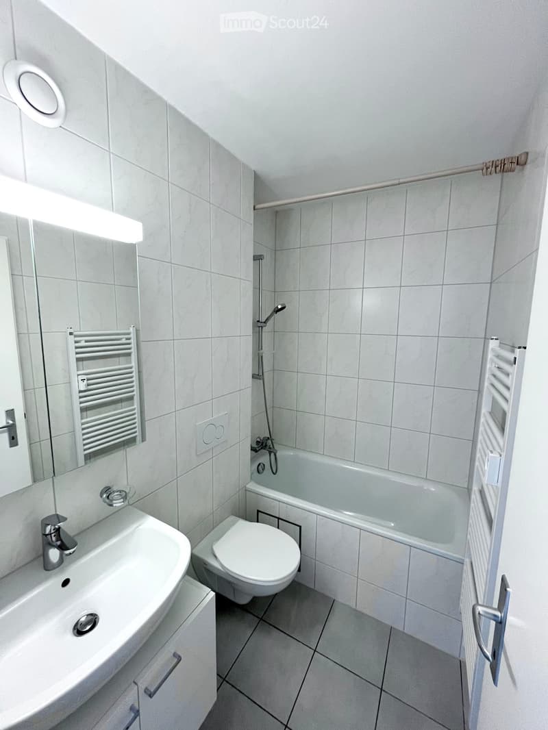 The image shows a bathroom with white tiles covering the walls and floor. It has a bathtub, a toilet, a sink, and a towel rack. The bathroom appears to be clean and well-maintained.