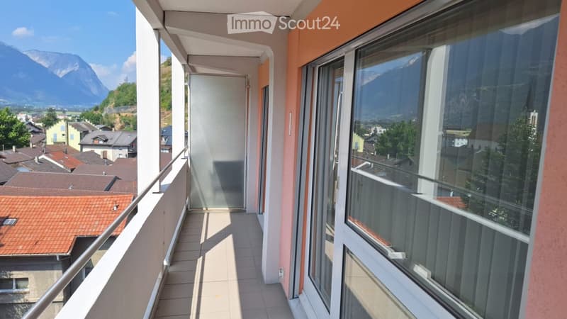 Balcony with tiled floor, metal railing, large windows offering a view of the town and mountains.