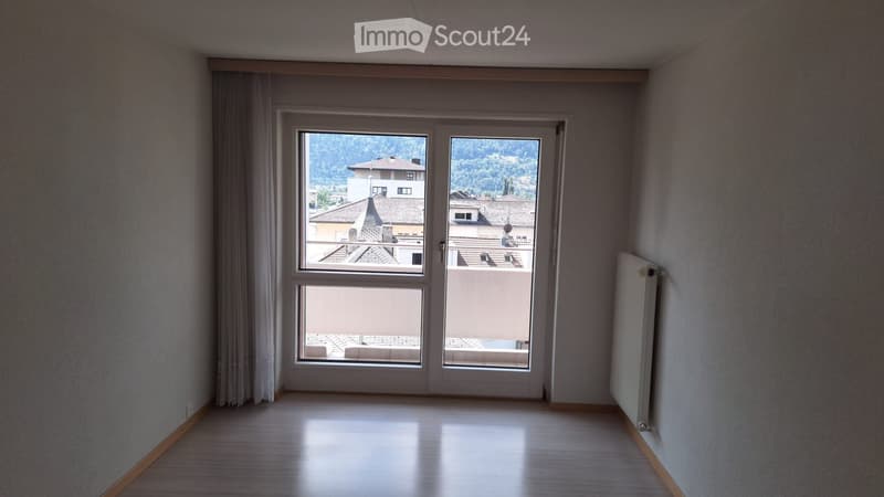 Empty room with wooden flooring, large window with view of rooftops and greenery, radiator on wall, and curtain partially drawn.
