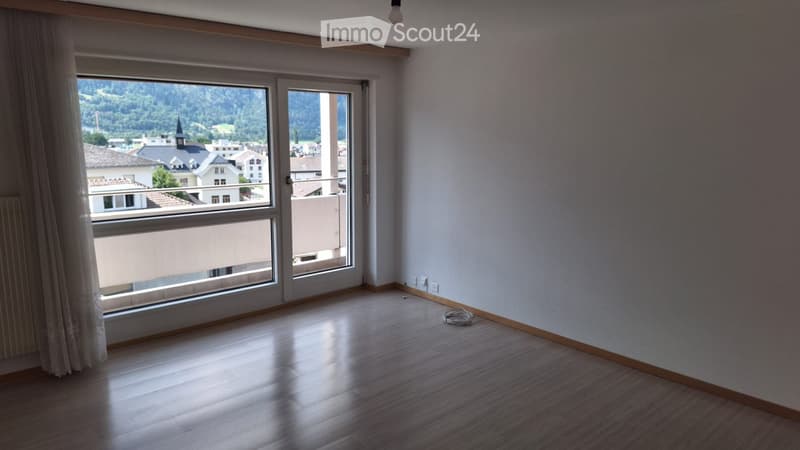 Empty room with wooden flooring, large windows, and a sliding door leading to a balcony. The room has white walls and a visible electrical outlet.