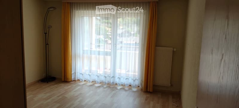 Empty room with wooden flooring, large window with sheer curtains, radiator, and a floor lamp near the window.