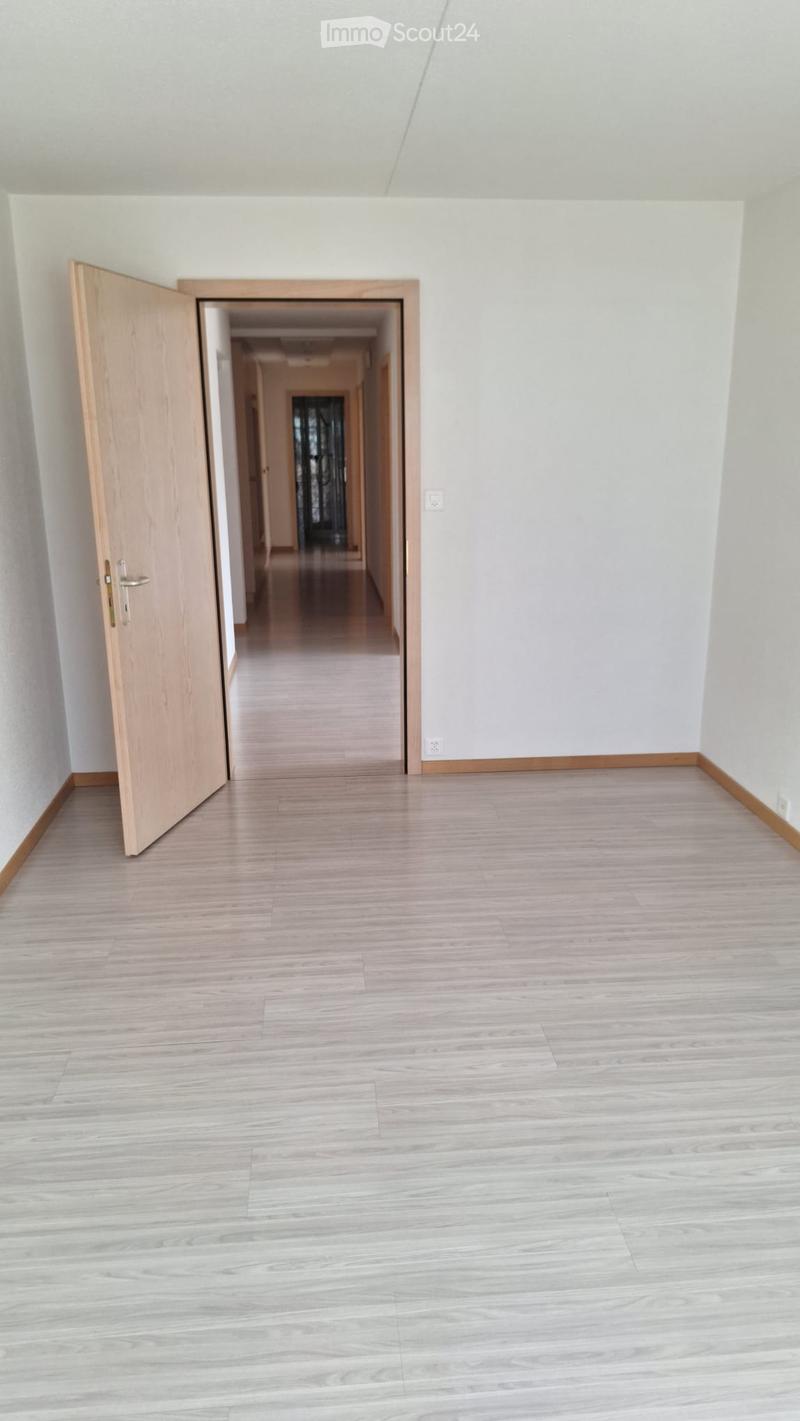 Empty hallway with light wooden flooring, white walls, and an open door leading to another area.
