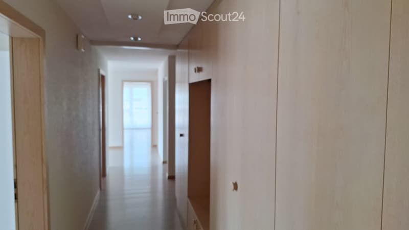A clean, modern hallway with white walls, wooden doors, and built-in storage cabinets. The floor is tiled, and recessed lighting is visible on the ceiling.
