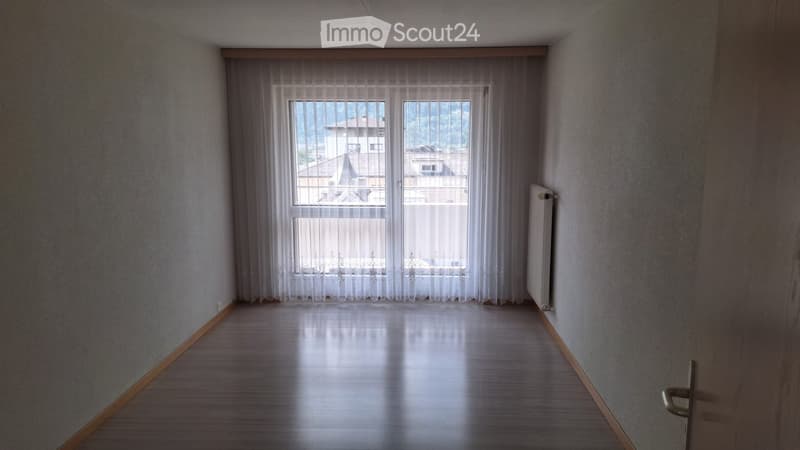 Empty hallway with wooden flooring, white walls, and large windows with sheer curtains, offering a view of rooftops and greenery outside.