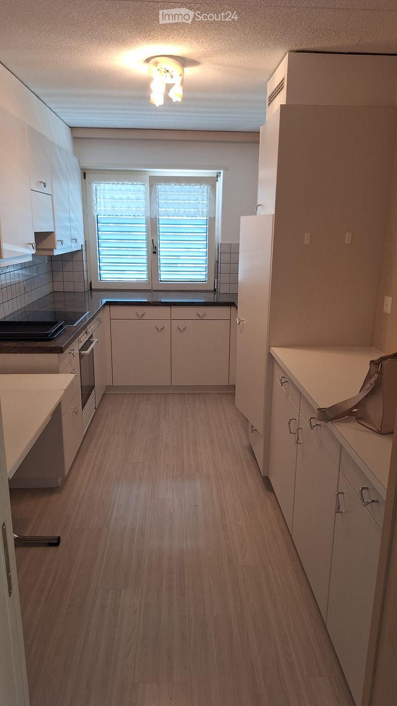 Modern kitchen with light wooden flooring, white cabinets, and a black countertop. Features an oven and a window with blinds.