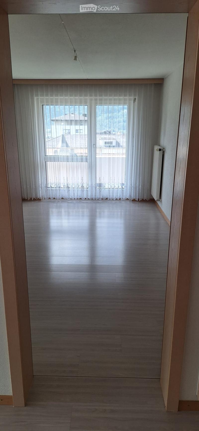 Empty room with wooden flooring, white walls, and large windows covered with sheer curtains. The room has a radiator on one wall and a visible ceiling light fixture.