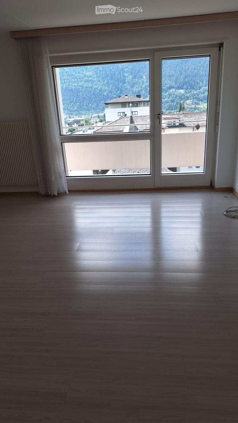 Empty room with wooden flooring, large windows, and a view of a forested area and neighboring buildings.