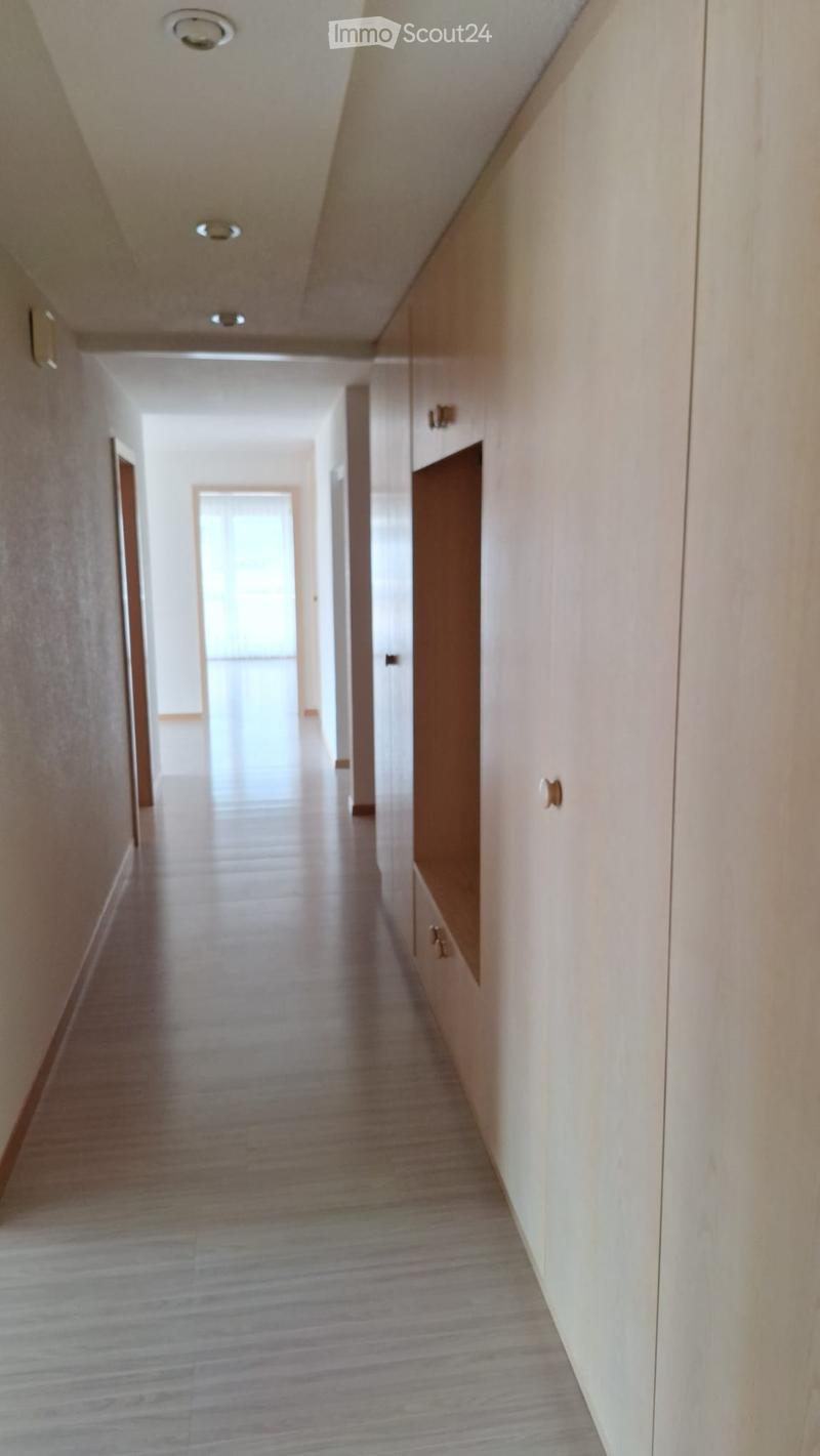 Clean, modern hallway with light wooden flooring, white walls, and built-in storage cabinets on one side. The hallway features recessed ceiling lighting and a visible doorway at the end.