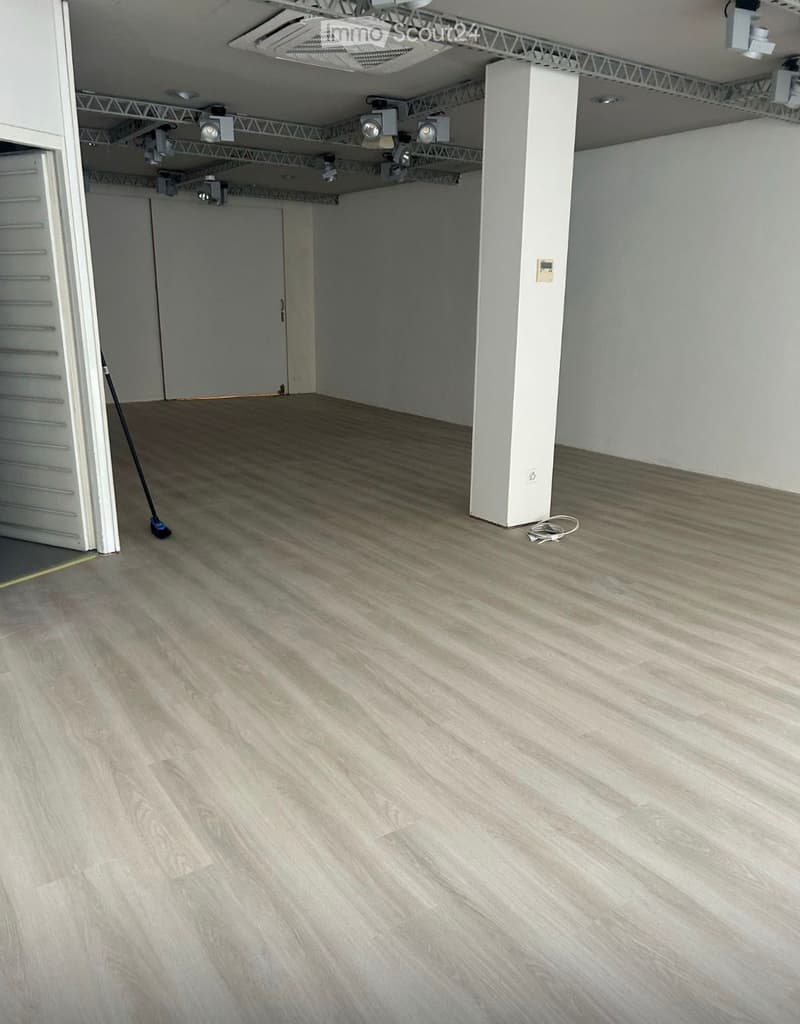 Empty room with light wooden flooring, white walls, and a single white pillar. A broom and a coiled cable are visible on the floor.