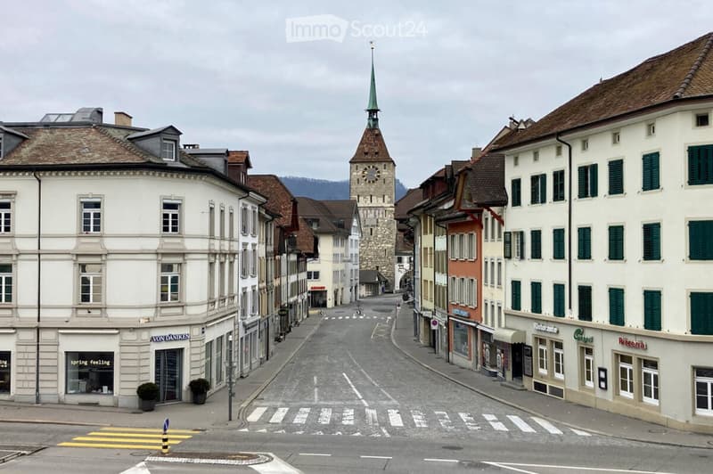 A quiet street lined with historic buildings, featuring a prominent clock tower in the background. The architecture includes a mix of white and colorful facades with green shutters, and the road is marked with pedestrian crossings.