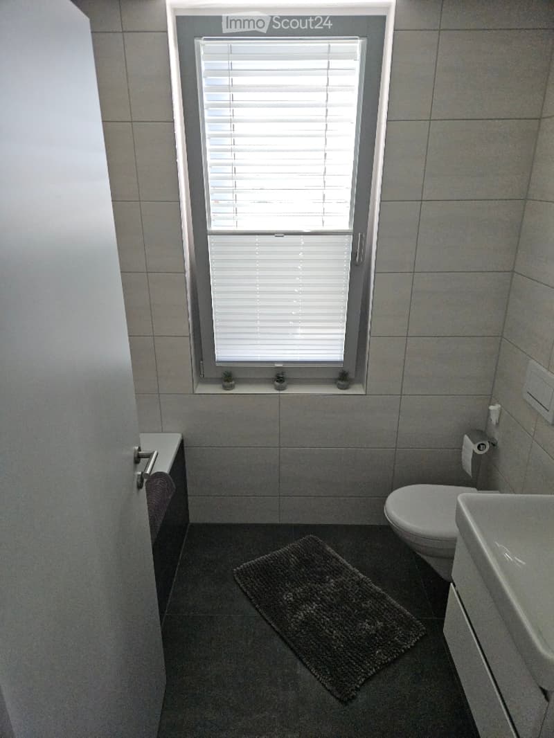 Modern bathroom with grey tiled walls and floor, a window with white blinds, a bathtub, a toilet, a sink, and a small bathroom rug on the floor.