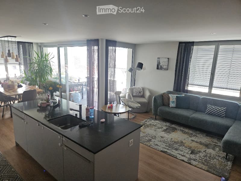 Modern open-plan living and dining area with a grey sectional sofa, coffee table, dining table with chairs, kitchen island with sink, and large windows leading to a balcony.
