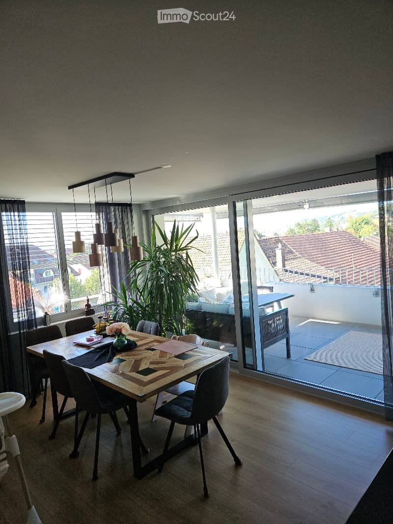 Modern dining area with a wooden dining table, six chairs, a large potted plant, and a view of a balcony or terrace through large windows. The room features contemporary decor, including a patterned table runner, a flower vase, and a candle holder.