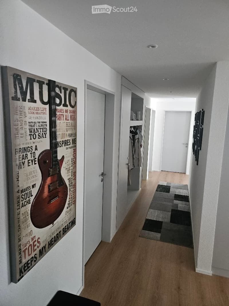 A modern hallway with light wooden flooring, white walls, and a large poster featuring a guitar and the word "MUSIC". A small rug is placed on the floor, and built-in shelving units are visible along the walls.