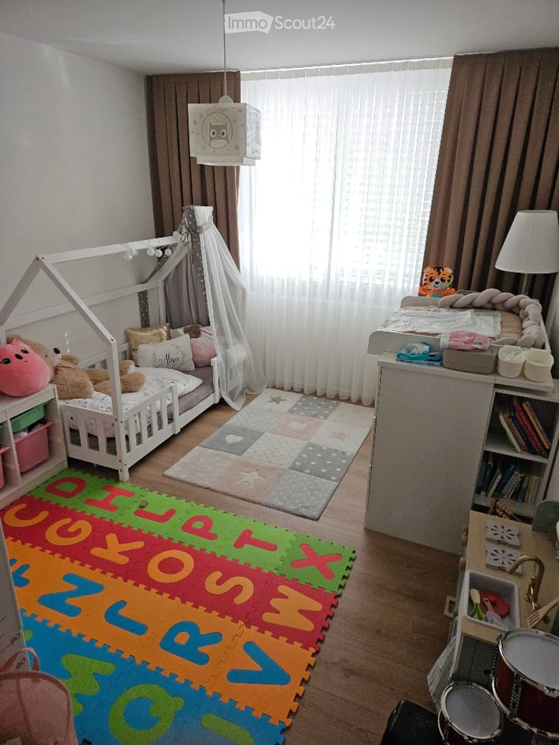 A well-lit children's bedroom with a white wooden bed, colorful alphabet play mat, shelves with toys, and a drum set in the corner.