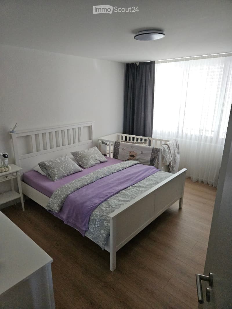 A cozy bedroom with a white wooden bed, purple bedding, a nightstand, and a baby crib in the corner. The room features wooden flooring and large windows with grey curtains.