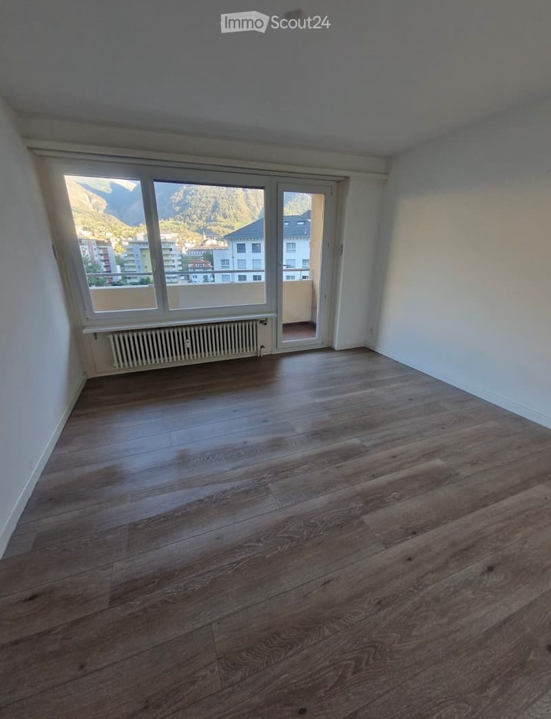 Empty room with wooden flooring, white walls, large window with view, and sliding glass door leading to a balcony.