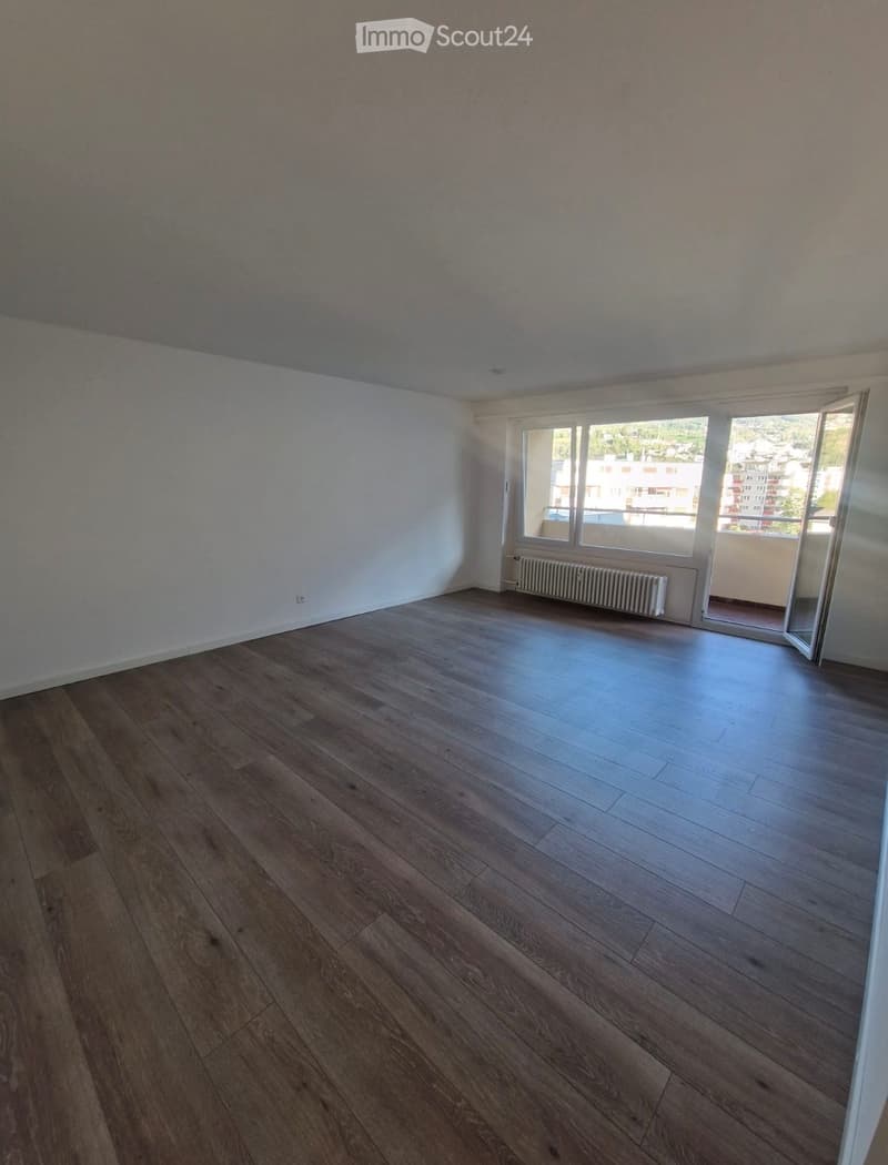 Empty room with wooden flooring, white walls, large window with view, radiator, and open sliding door leading to balcony.