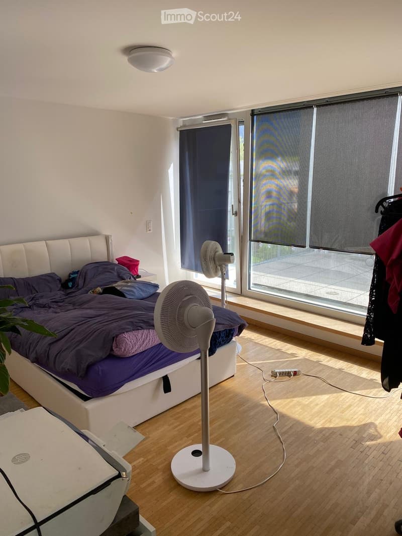 A modern bedroom with a white upholstered bed, purple bedding, a standing fan, a laptop on a glass table, and large windows with roller shades.