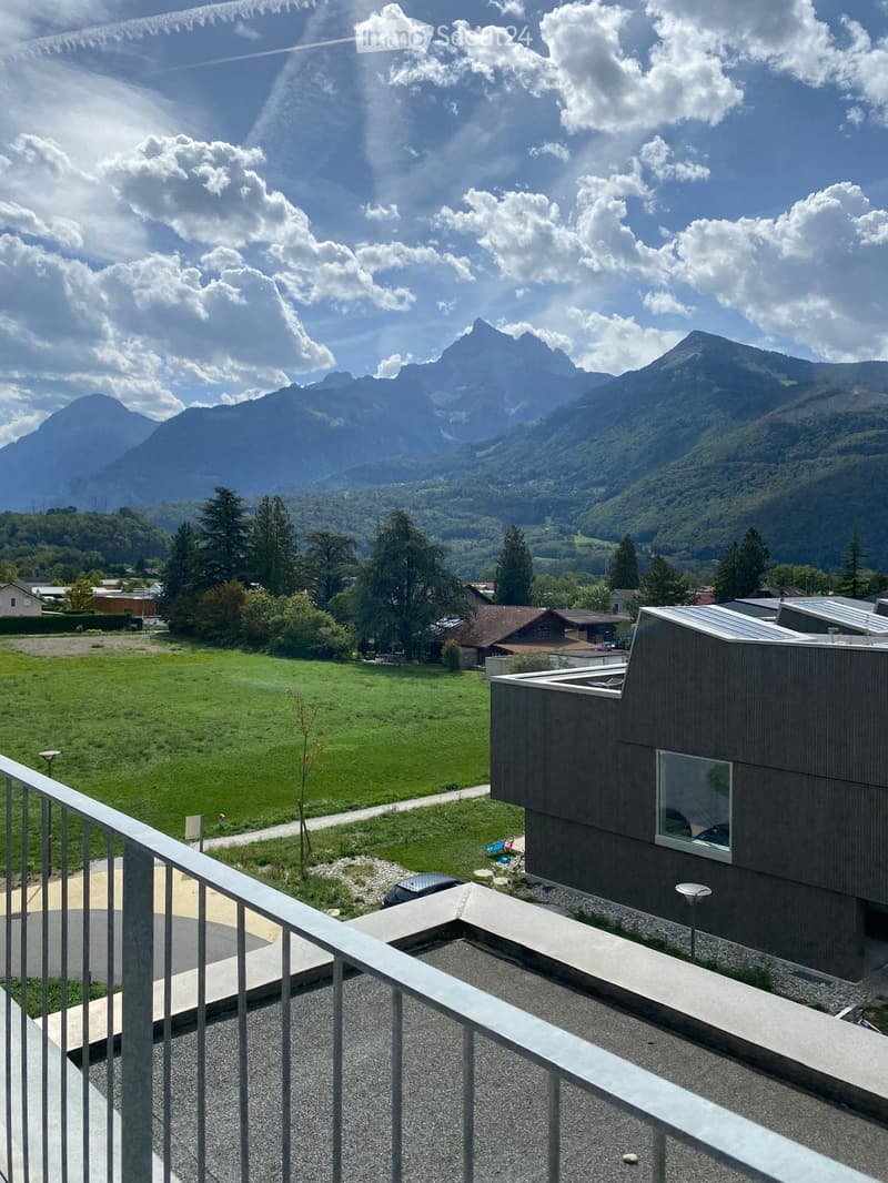 Balcony with a modern railing, overlooking a green field, scattered trees, and distant mountains under a partly cloudy sky.