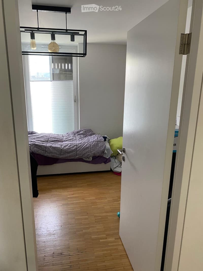 A small, well-lit bedroom with wooden flooring, a bed with a grey duvet and pillows, and a modern pendant light fixture above.
