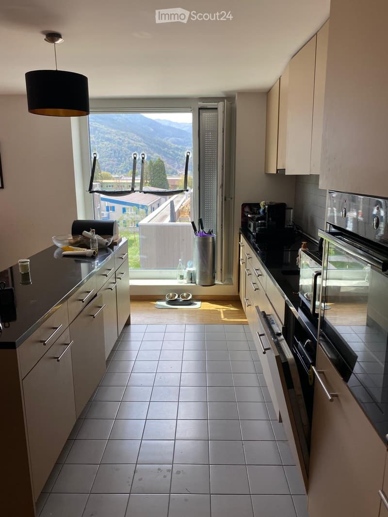 Modern kitchen with light-colored cabinets, tiled floor, and a large window offering a scenic mountain view. Countertop holds a bowl, bottle, and other small items.