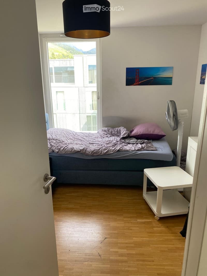 Cozy bedroom with wooden flooring, a double bed with grey bedding, a white nightstand, a fan, and a small white table near the door.