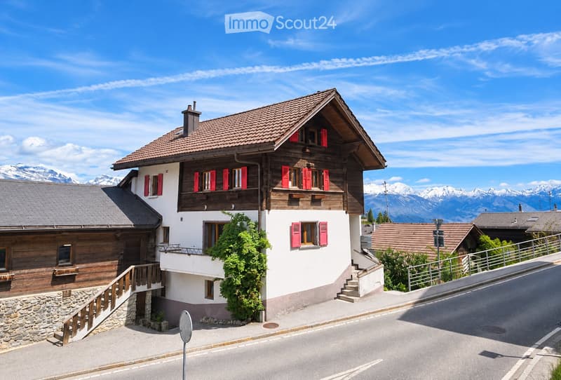 A two-story house with a wooden facade, red shutters, and a stone base, situated in a scenic mountainous area with a clear blue sky and distant mountain peaks.