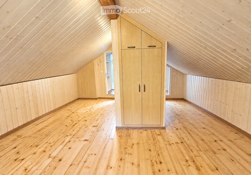 Empty attic space with wooden walls, ceiling beams, and hardwood flooring. Features built-in wooden cabinetry and large windows allowing natural light.