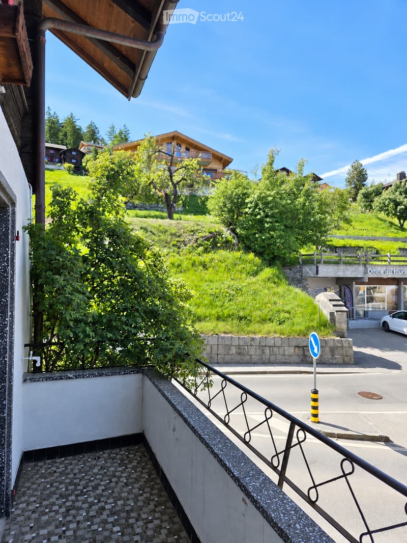 Balcony overlooking a green hillside with trees and a wooden house in the background, featuring a metal railing and tiled steps.