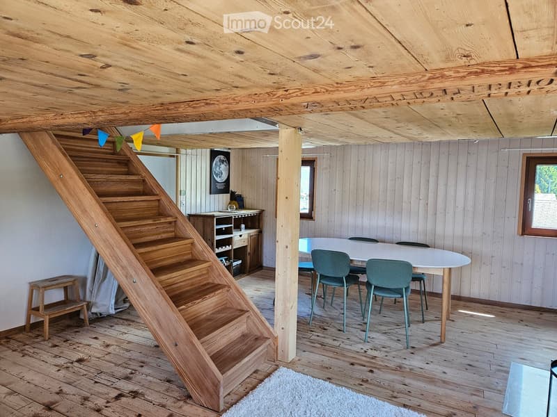 Furnished Room. Cozy wooden interior with a staircase leading upwards, a dining table with chairs, a small cabinet, and a wooden stool near the staircase.