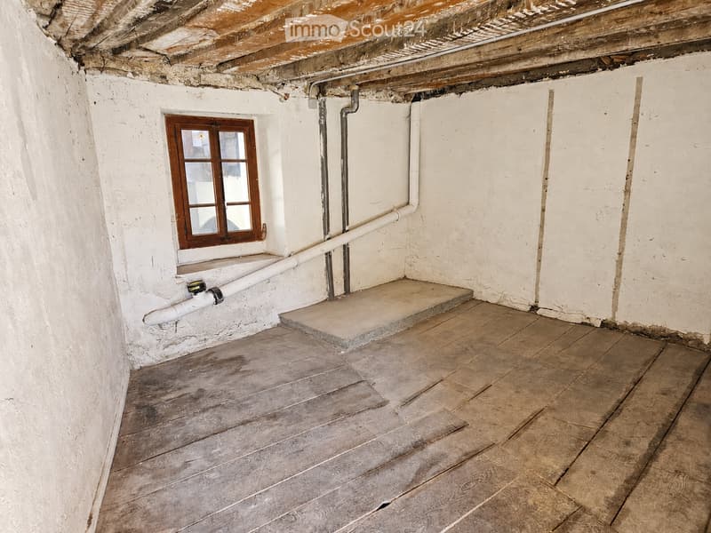 Empty room with wooden floor, white walls, exposed ceiling beams, and a small window with wooden frame. Exposed pipes and a small step leading up to a platform are visible.