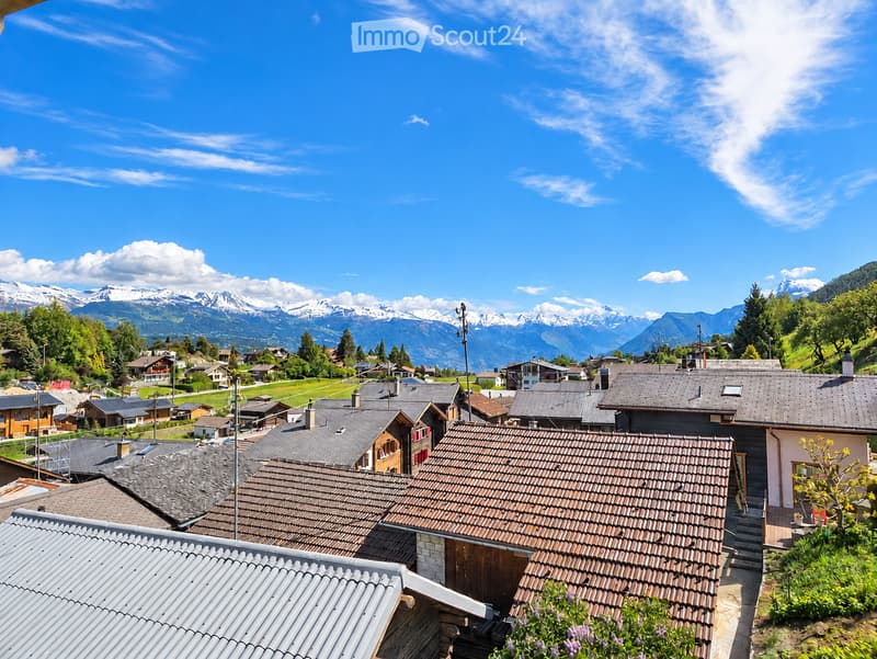 A panoramic view of a village with traditional houses and tiled roofs, surrounded by lush greenery and snow-capped mountains under a clear blue sky.