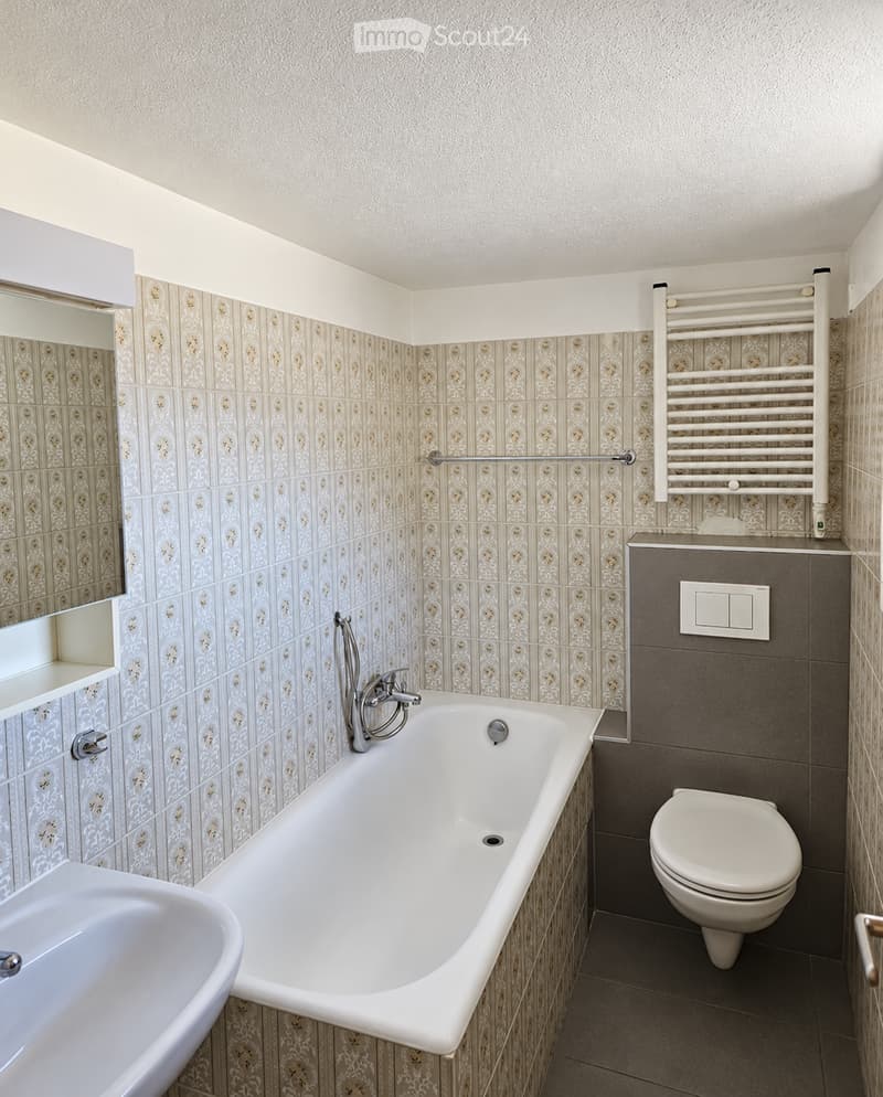 Bathroom with a bathtub, toilet, sink, mirror, and a radiator. The walls are tiled with a decorative pattern, and the floor is tiled in a neutral tone.