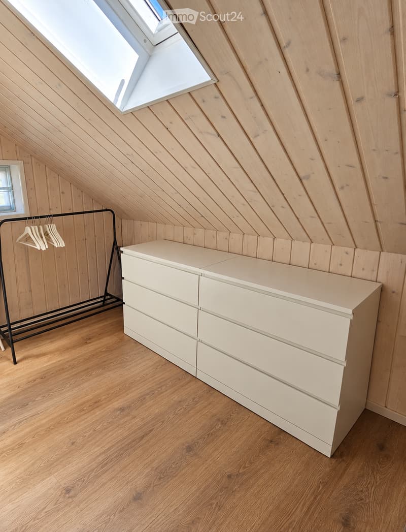 Attic room with wooden walls and floor, skylight window, white dresser, and black clothing rack with hangers.