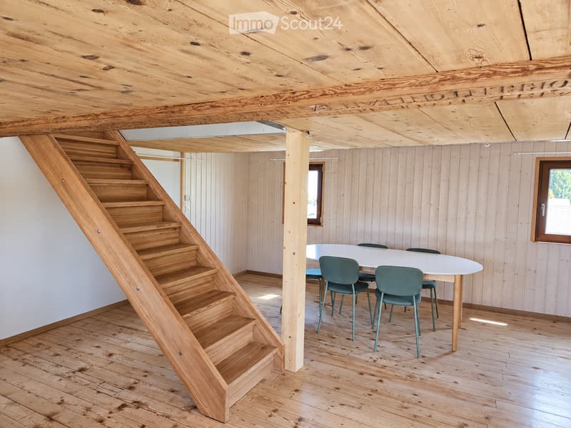 Wooden staircase leading up, white table with four green chairs, wooden walls and ceiling, natural light from windows.