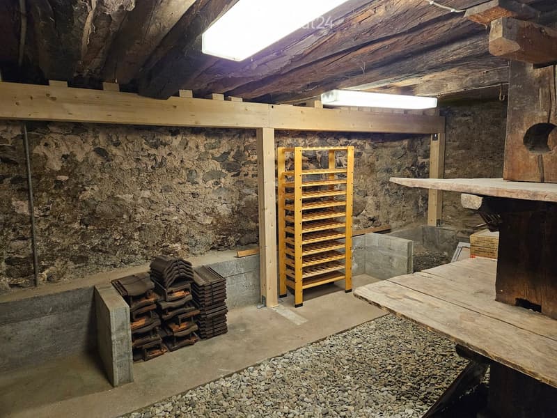 Unfurnished Room. An attic or storage space with exposed wooden beams, stone walls, and a concrete floor. Contains a wooden shelving unit, a stack of metal items, and a wooden table.