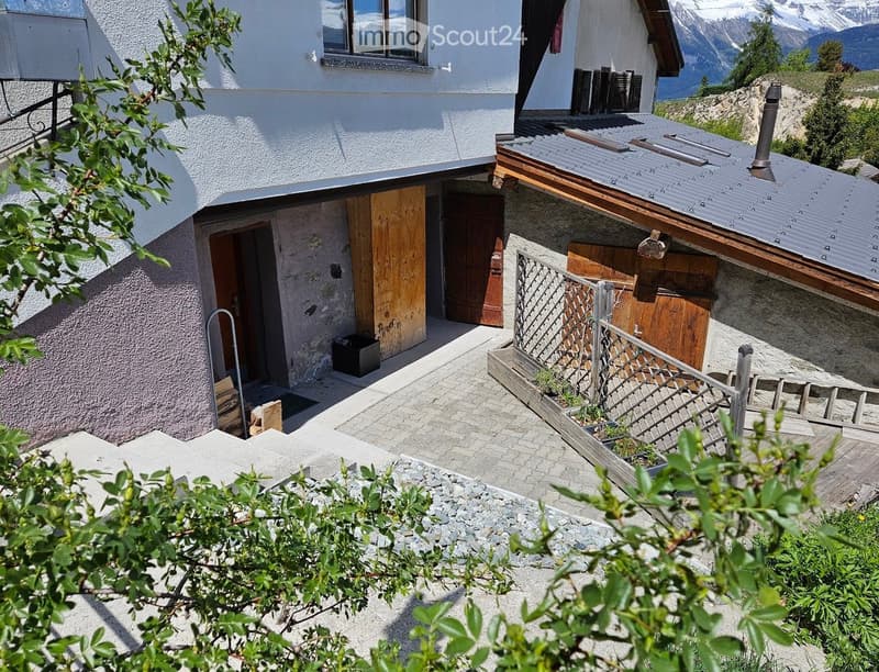 Building. A two-story white building with wooden doors and windows, situated in a mountainous area with a view of snow-capped peaks. The entrance features a small porch with a metal railing and some stacked wood.