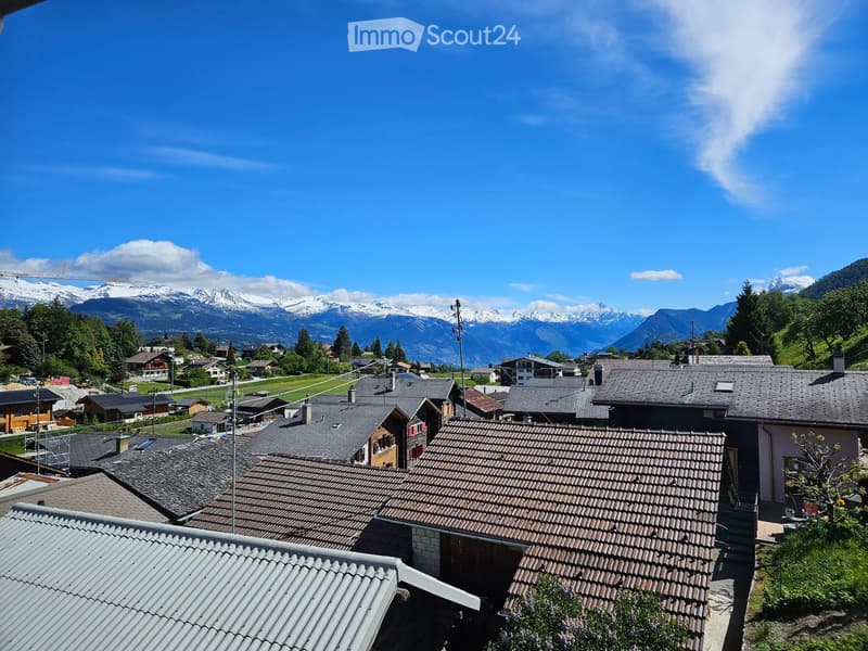 Building. A panoramic view of a village with traditional houses, tiled roofs, and a backdrop of snow-capped mountains under a clear blue sky.