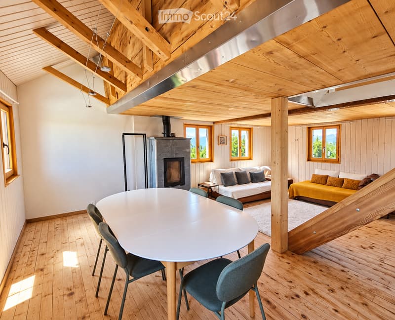 A spacious, well-lit room with wooden beams and exposed wooden ceiling. Features include a round dining table with chairs, a sofa, a bed with pillows and blankets, a fireplace, and large windows with scenic views.