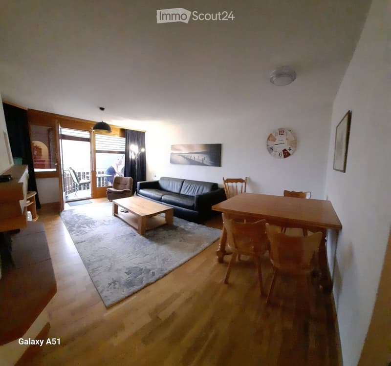 Living room with a black leather sofa, wooden coffee table, dining table with chairs, a large clock on the wall, and a picture frame. The room has wooden flooring and a large rug.