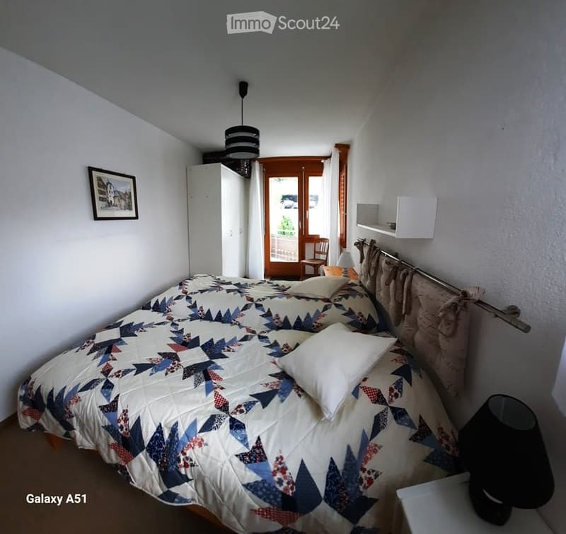 A cozy bedroom with a double bed featuring a patterned quilt and pillows, a wardrobe, a picture frame on the wall, a chair near the door, and a lamp on a nightstand.