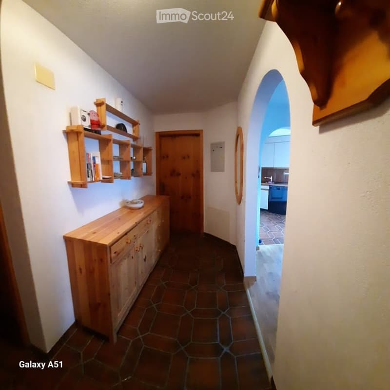 A narrow hallway with wooden shelves on one side, a wooden cabinet, and a door leading to another room. The floor is tiled, and the walls are painted white.