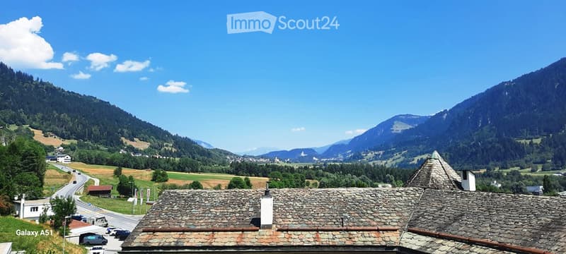 A picturesque view of a traditional stone-roofed building nestled in a lush, green valley surrounded by towering mountains under a clear blue sky.