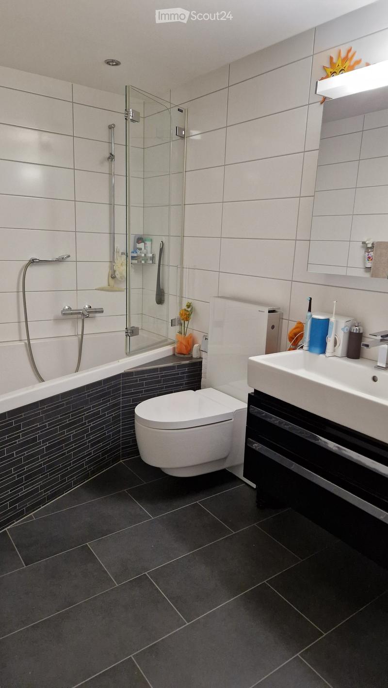 Modern bathroom with dark tiled floor, white bathtub, toilet, and sink. Features a glass-enclosed shower area with a handheld showerhead, and a shelf with toiletries and a small flower vase.