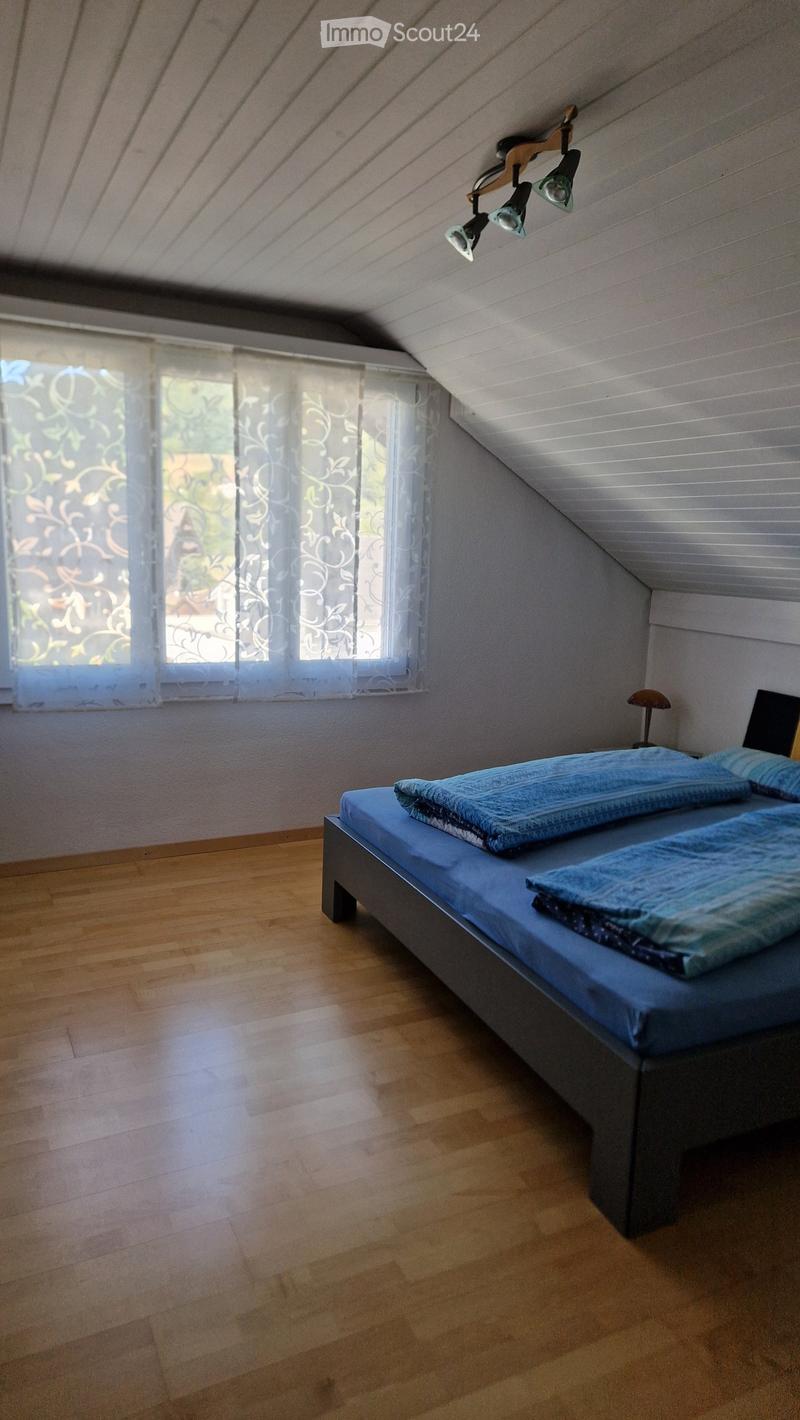 Bright bedroom with wooden floor, large window with sheer curtains, bed with blue bedding, and a small lamp on a nightstand.