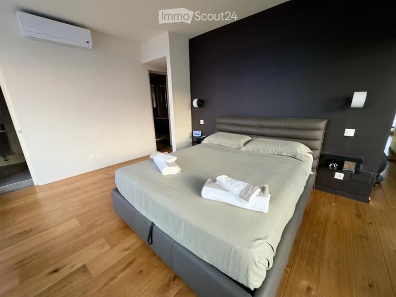 Modern bedroom with a large bed, grey bedding, wooden flooring, and a dark accent wall. Includes a nightstand and a mirror.